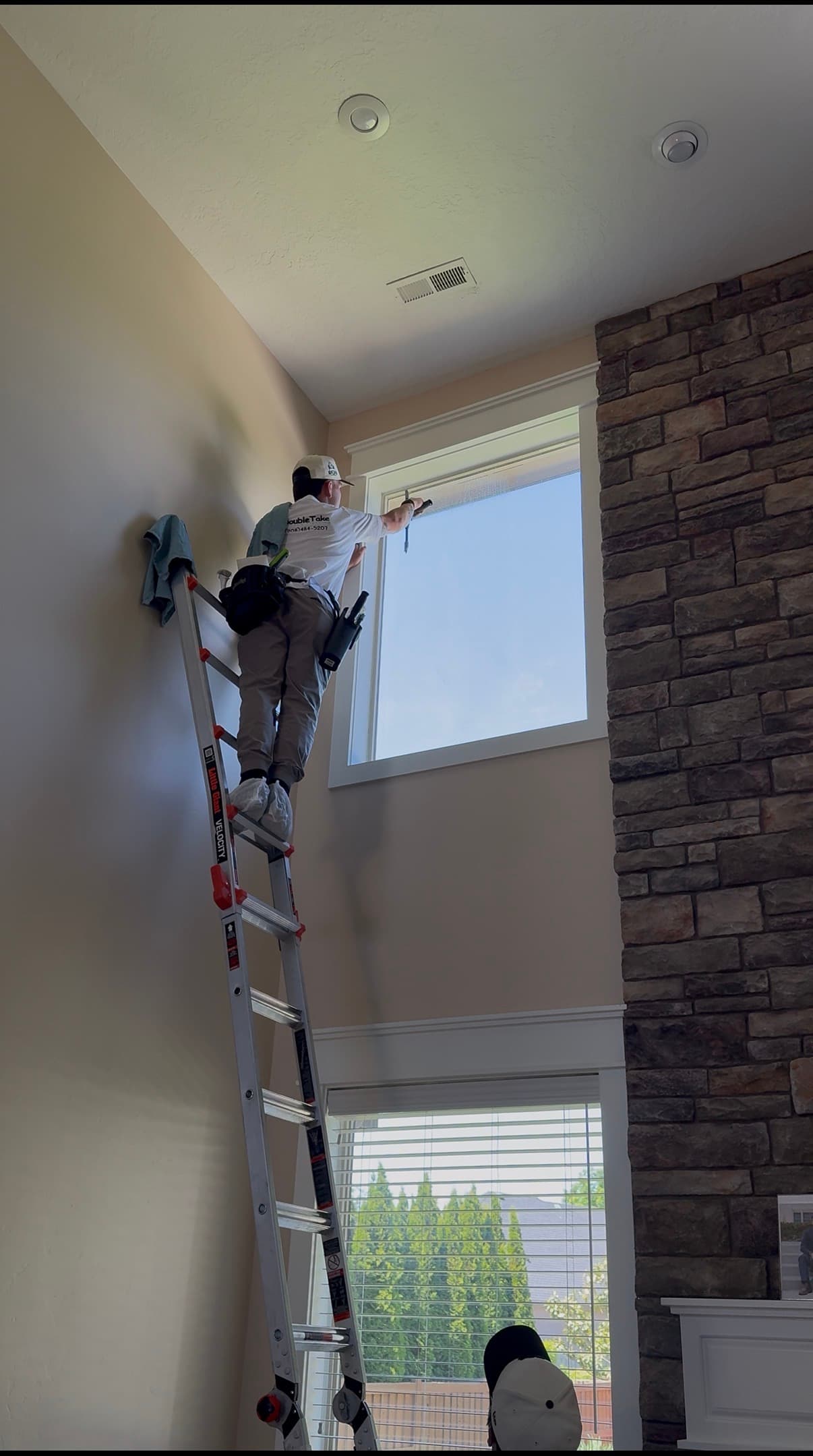 Professional window cleaner on a tall ladder cleaning a high window next to stone fireplace.