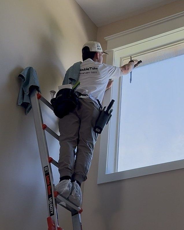 A professional window cleaner on a ladder uses a squeegee on a high indoor window.